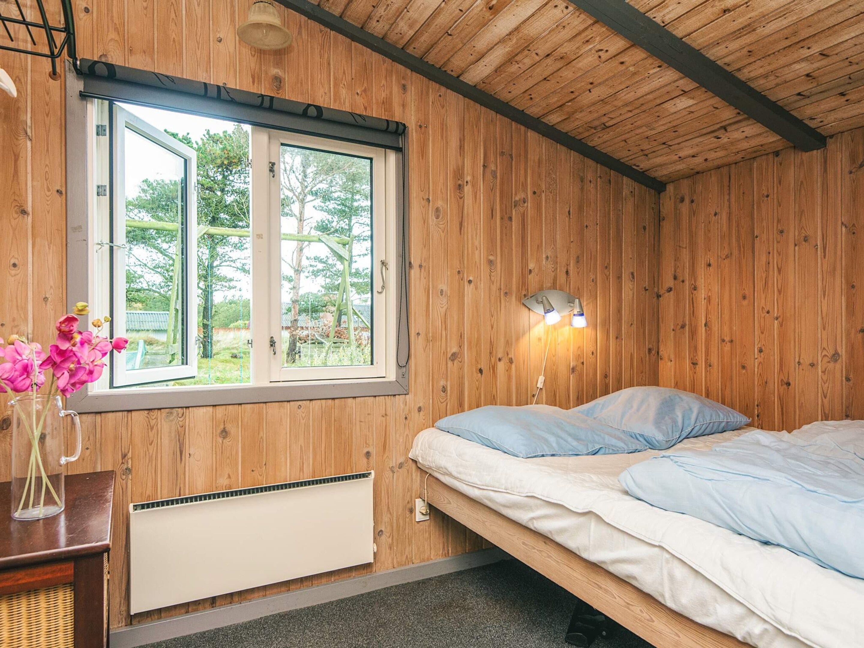 Una habitación con paredes de madera, una cama con ropa de cama azul, una ventana que da a zonas verdes y una lámpara fijada a la pared.