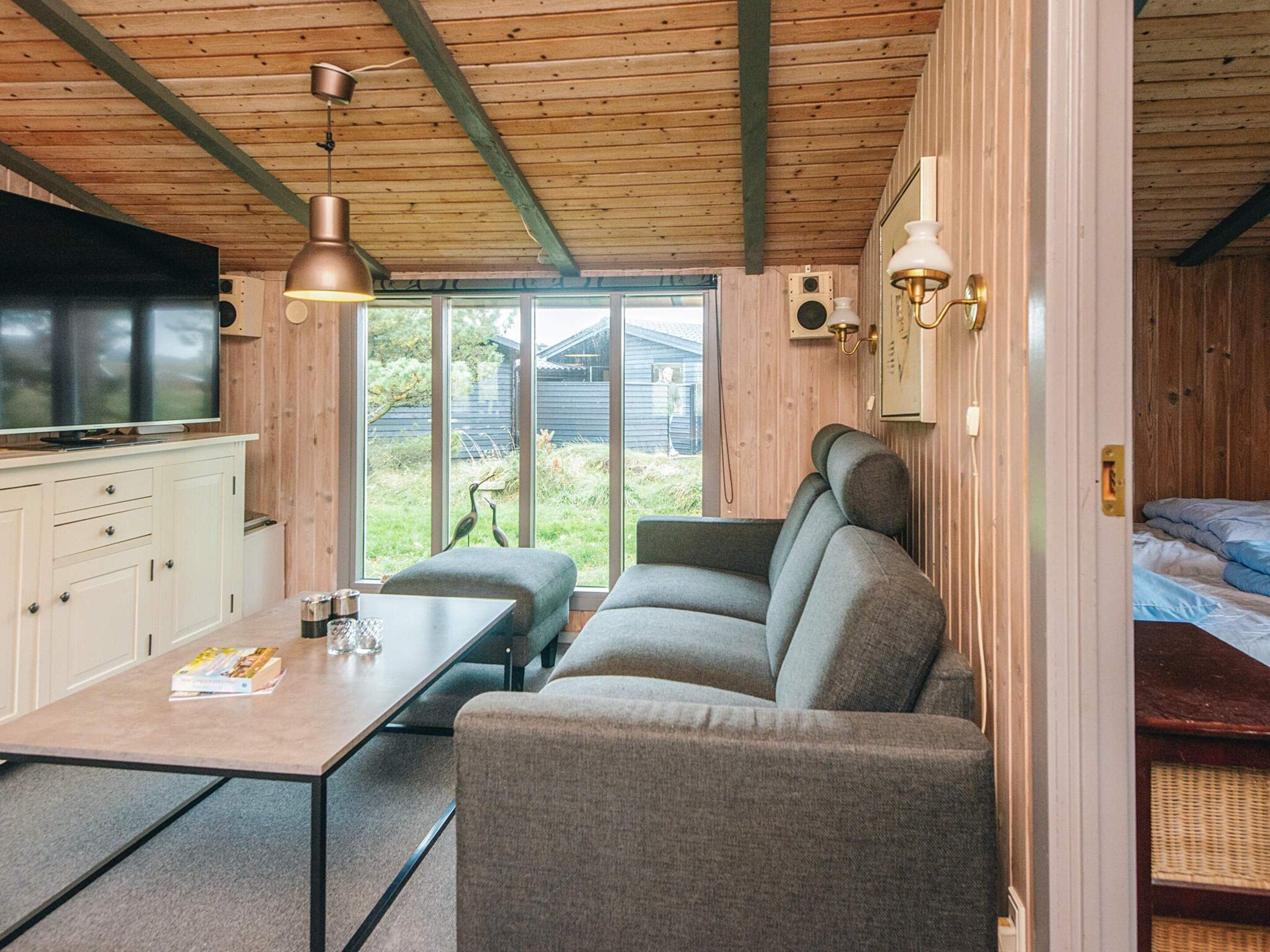 A cozy room with a sofa, a table, a television, and a window with a view of greenery.