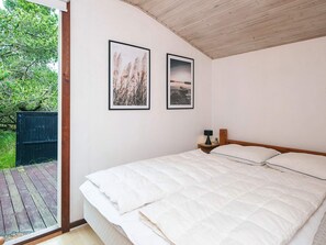 Une chambre avec un plafond en bois, un lit recouvert de linge de lit blanc, deux tableaux encadrés au mur et une vue sur une terrasse en bois et la verdure à l’extérieur.