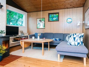 Un salon chaleureux avec un plafond en bois, une grande fenêtre donnant sur les arbres, un canapé gris, une petite table et une cheminée.