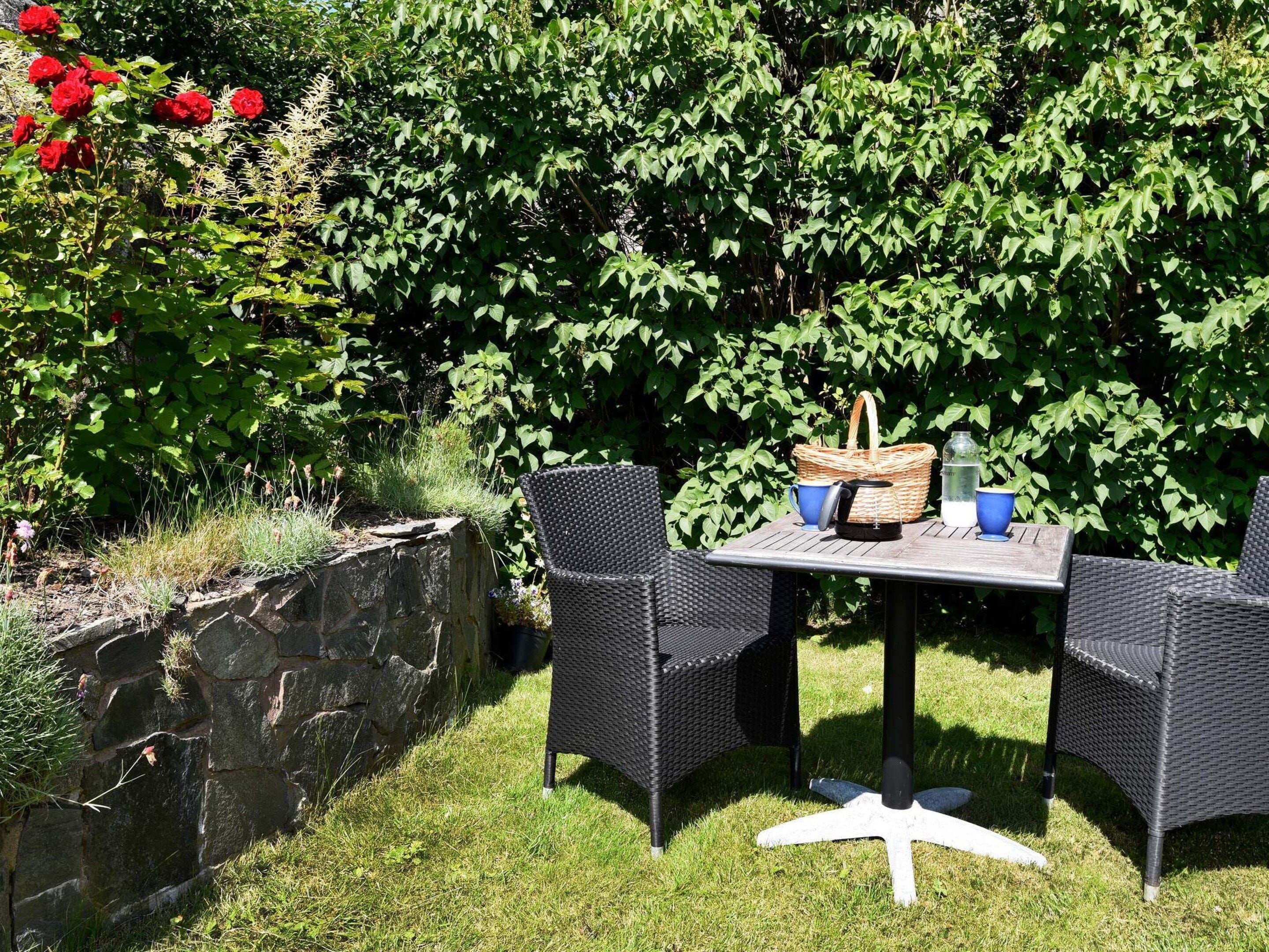 An outdoor seating area with wicker chairs and a table, surrounded by greenery and a stone wall.