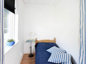 Quarto pequeno com uma cama de solteiro, cabeceira de madeira, mesa de cabeceira com luminária, um vaso de planta no parapeito da janela e um travesseiro listrado.