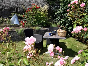 An outdoor seating area with wicker chairs and a table, surrounded by lush greenery and flowering plants. - Holiday Home in Skærhamn (Skaerhamn)