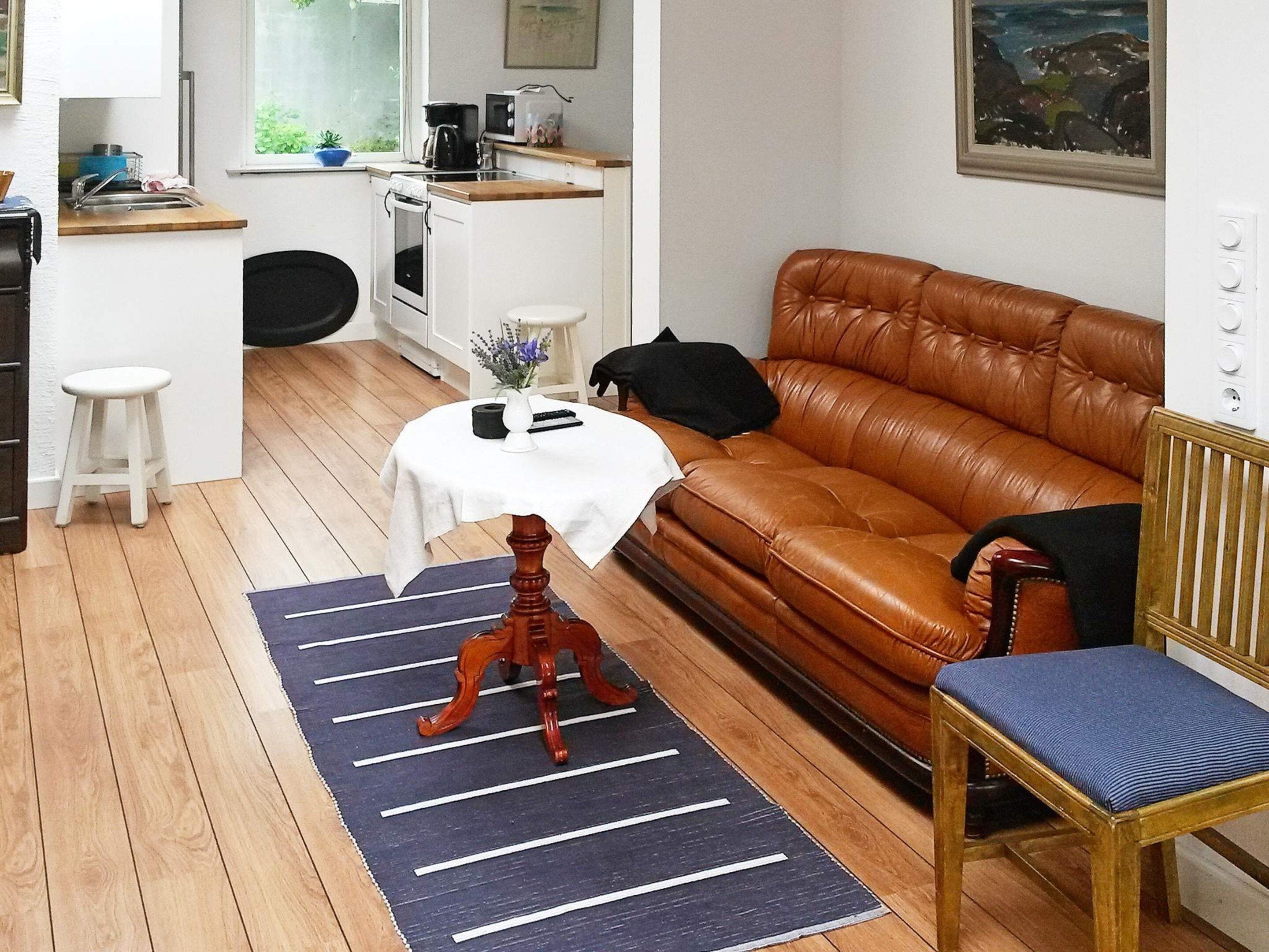 Ett kompakt vardagsrum med en brun lädersoffa, ett litet runt bord med en vit duk, en trästol och ett köksområde i bakgrunden.
