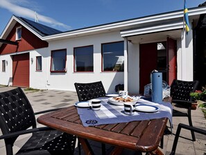 A wooden table with chairs, a plate of food, and a mug on a checkered tablecloth outside a building with windows and a red door. - Holiday Home in Skærhamn (Skaerhamn)