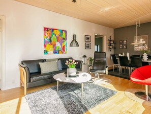 Ein modernes Wohnzimmer mit einer schwarzen Ledersofa, einem runden Couchtisch mit Blumen in einer Vase, einem farbenfrohen abstrakten Gemälde an der Wand und einem Essbereich mit einem roten Stuhl.