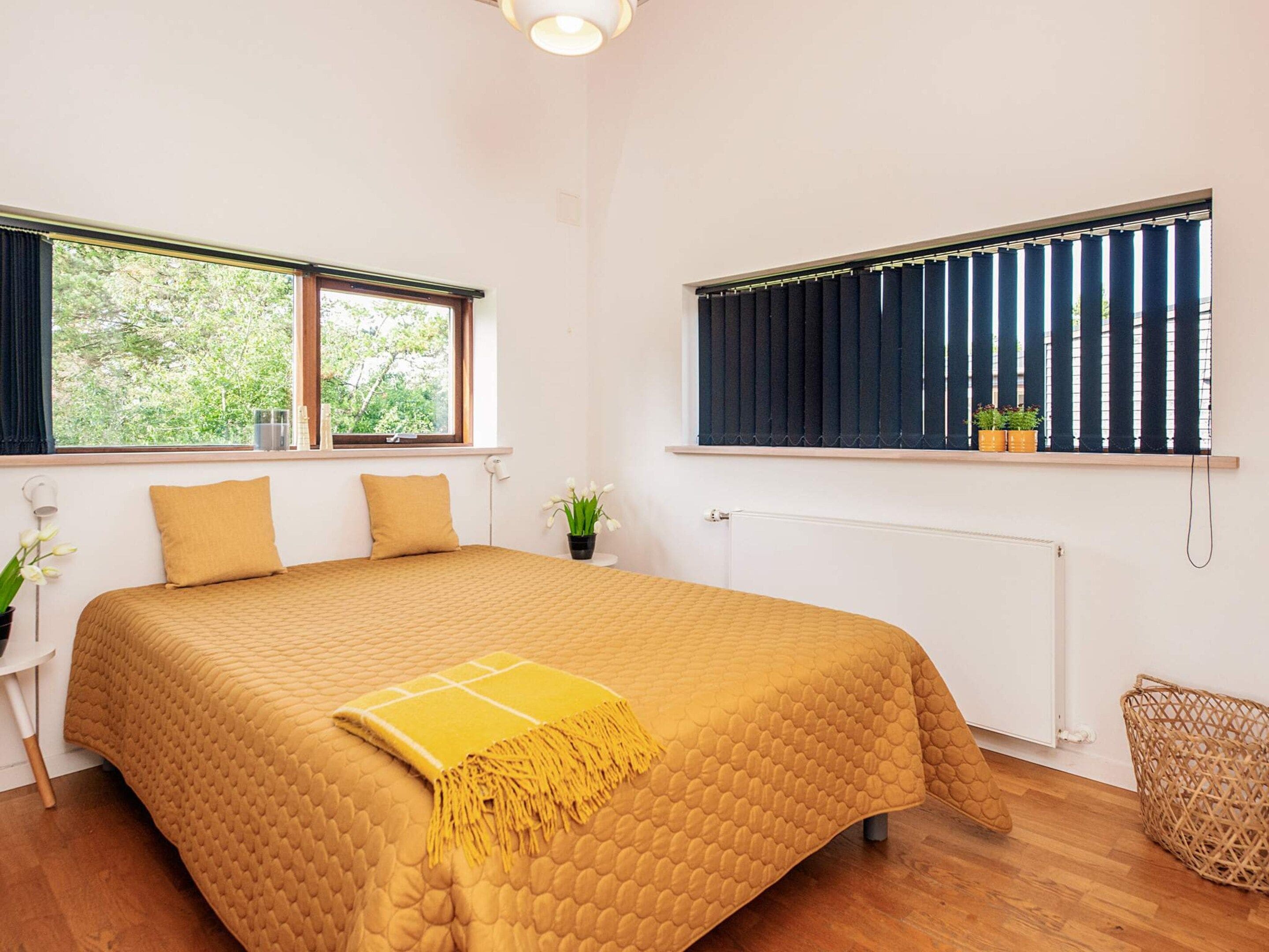 Quarto com cama, duas janelas, radiador, mesa de cabeceira com abajur e planta em vaso.