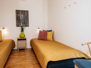 A room with two beds, a wooden floor, a wall-mounted painting, a small table with a green vase, and a chair.