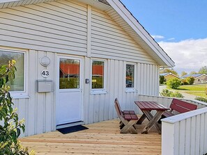 Un bâtiment d’un seul étage, à l’extérieur blanc, avec une porte d’entrée et un petit porche comprenant une table en bois et deux chaises.