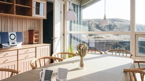 Zona pranzo con tavolo apparecchiato per due, sedie di legno, un vaso con fiori secchi e vista su un paesaggio montuoso attraverso ampie finestre.