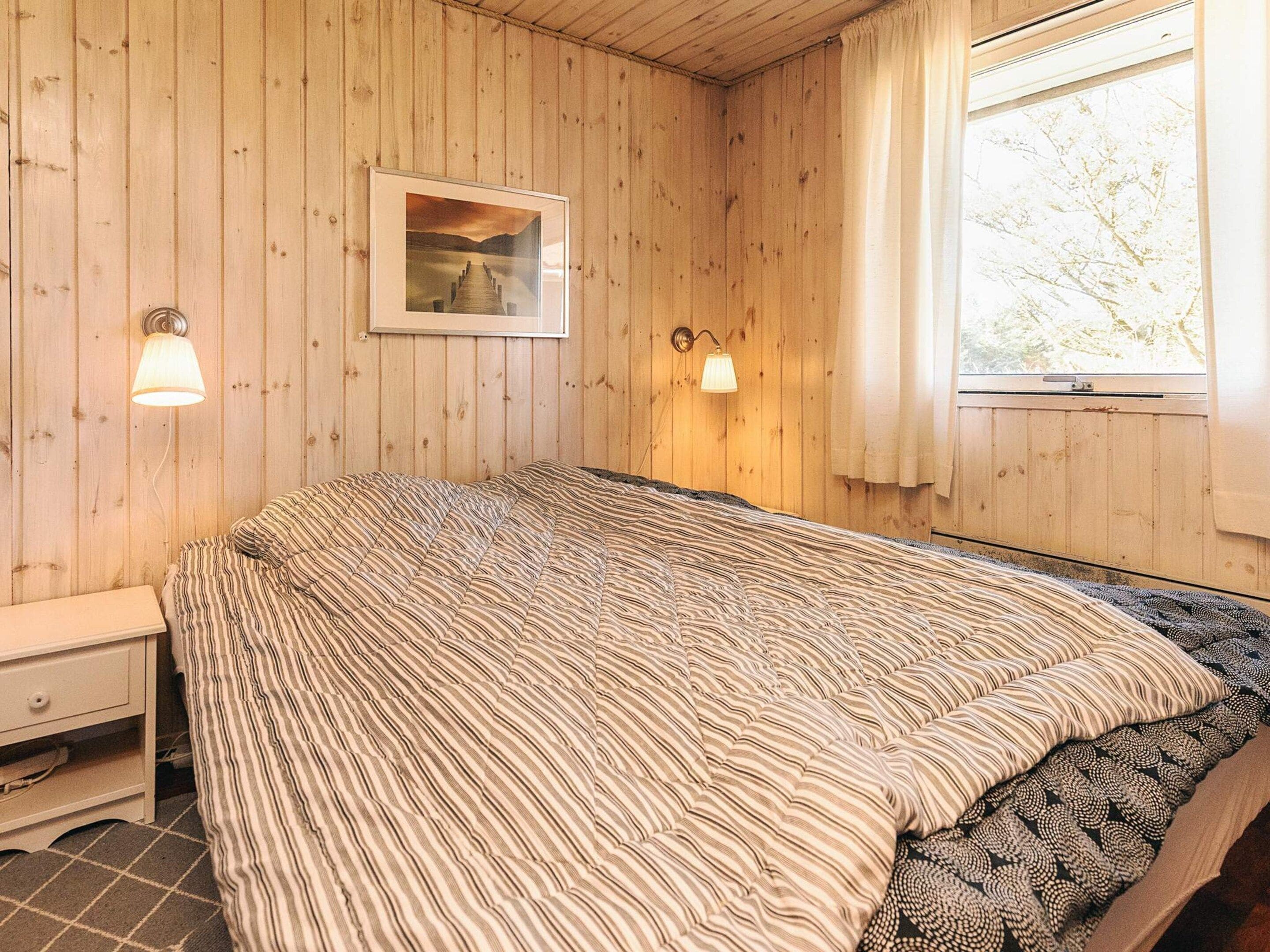 Quarto com painéis de madeira, cama, mesa de cabeceira, abajur e um quadro na parede.