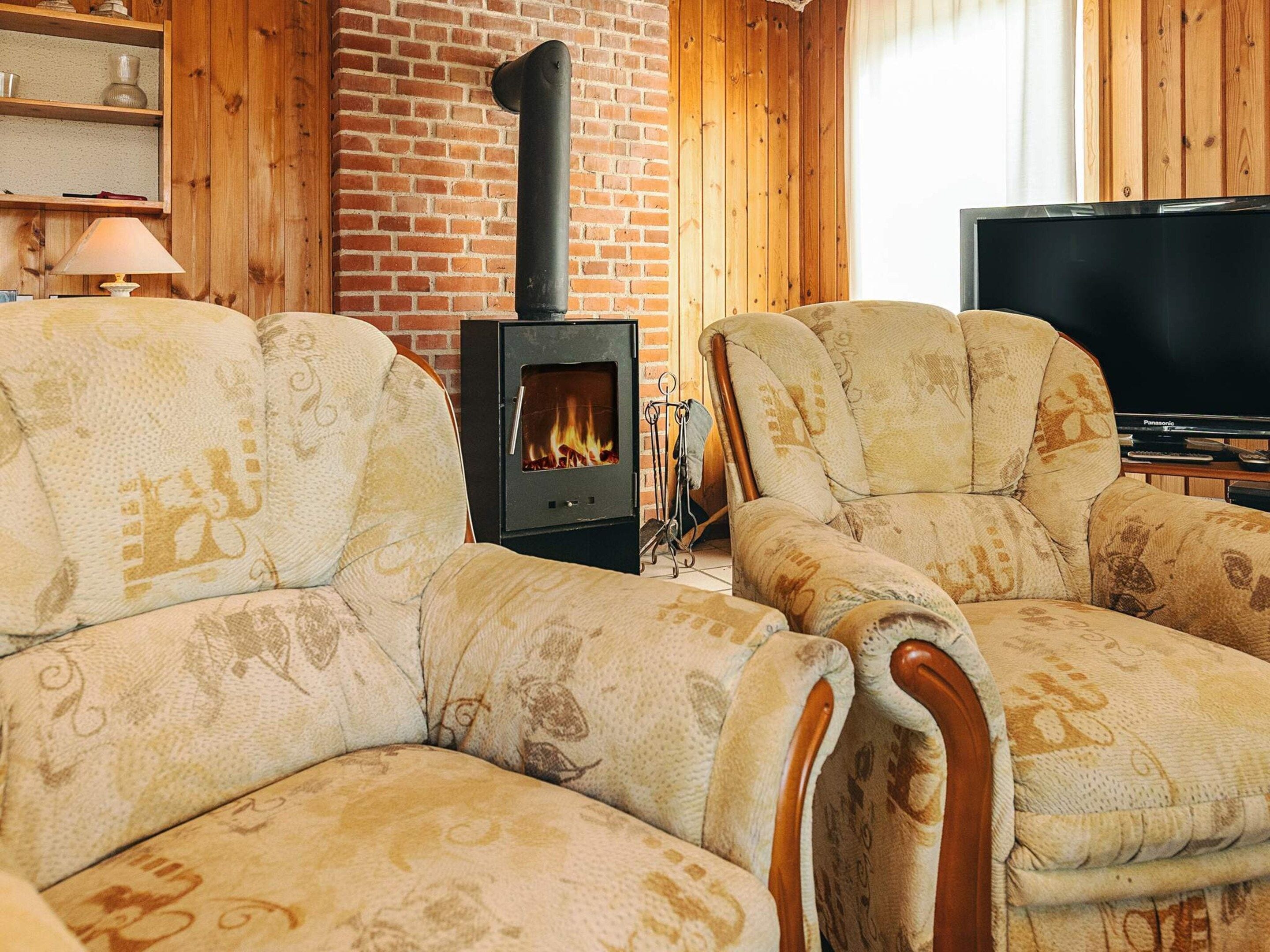A cozy living room with two patterned armchairs, a brick wall, a wood-burning stove, and a television.