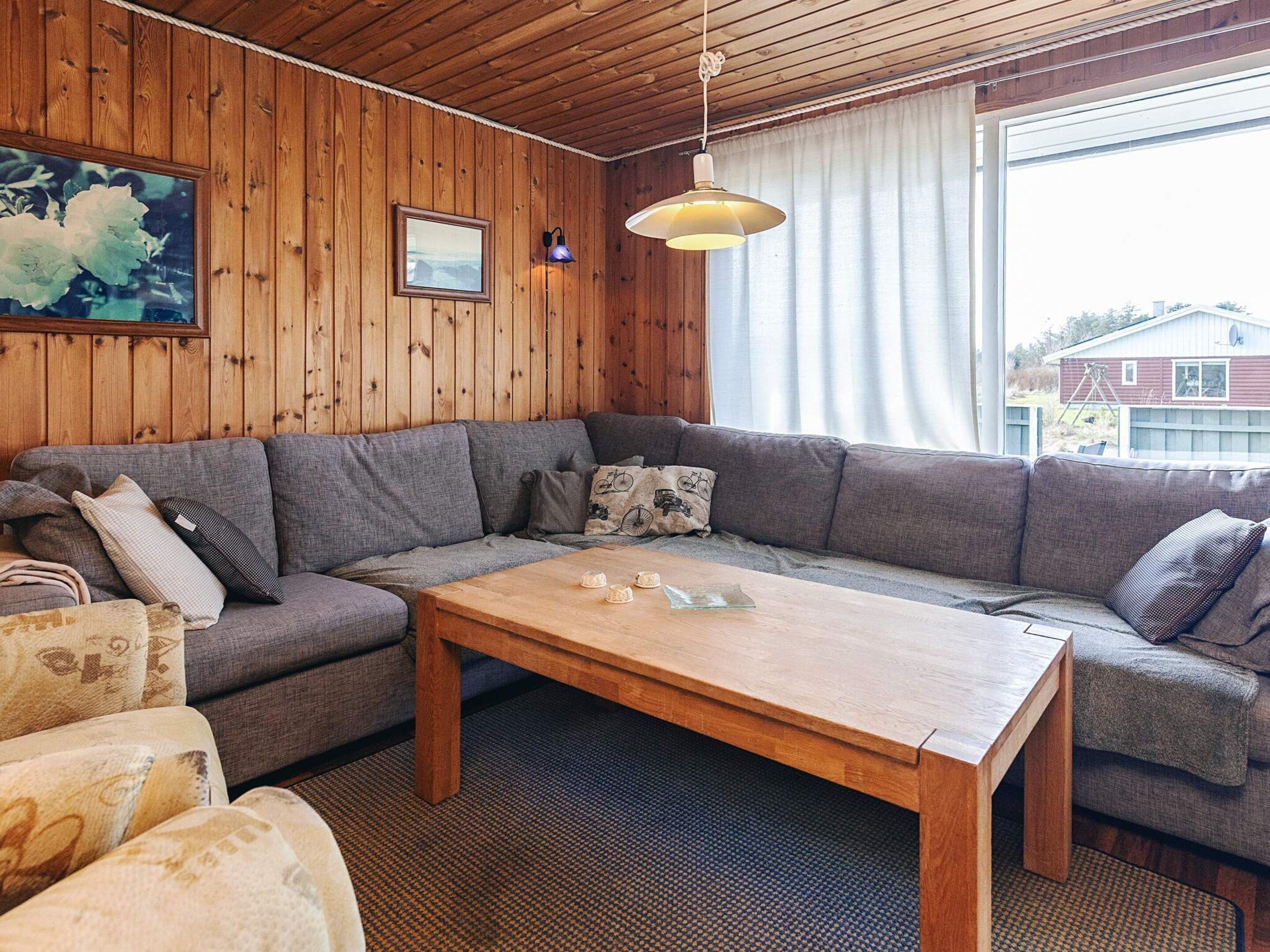 Ett mysigt vardagsrum med ett träpanelat tak, en grå soffa, ett träbord och ett stort fönster med utsikt över ett hus.