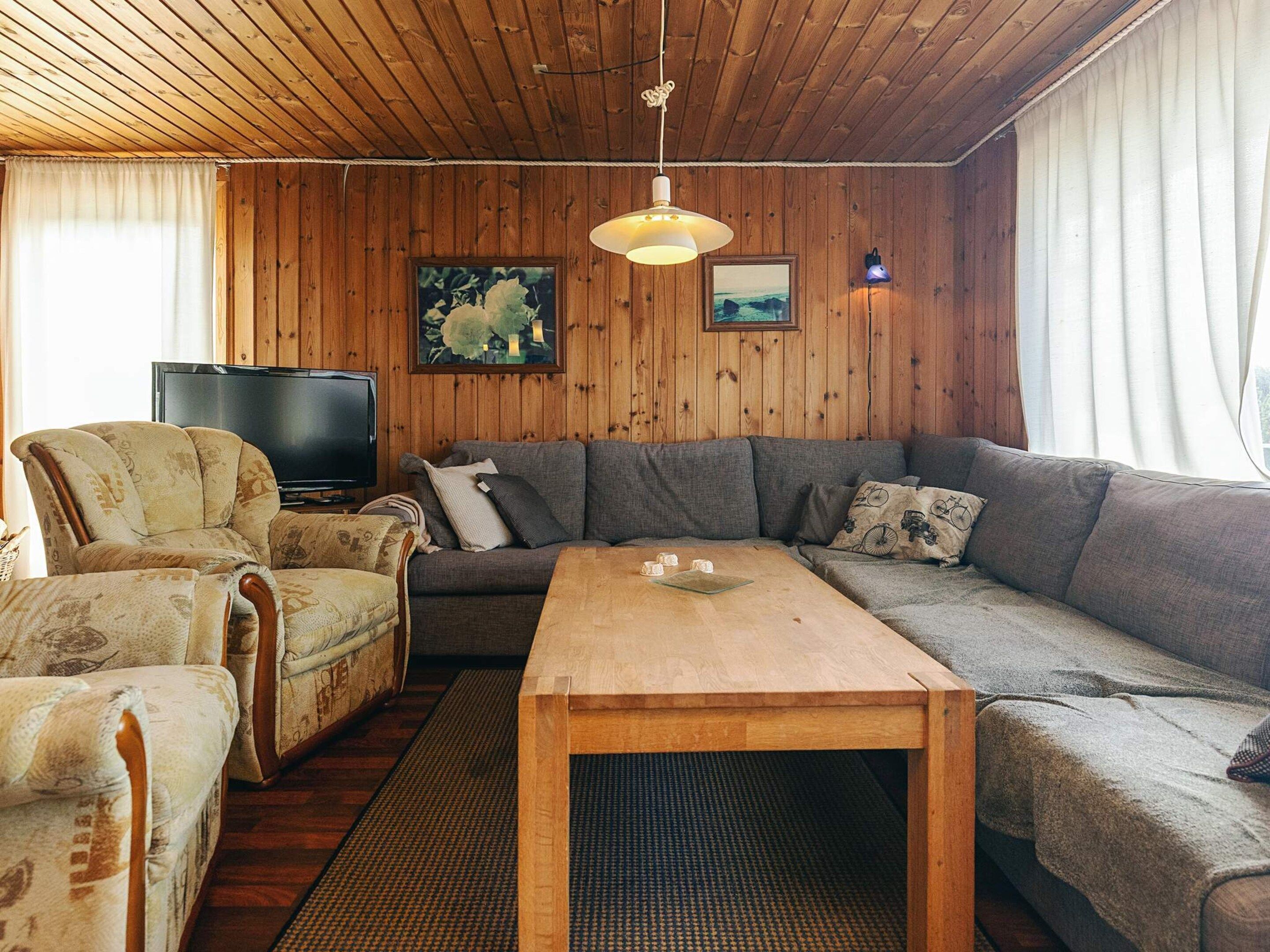 A cozy living room with a wooden ceiling, a large grey sofa, a wooden coffee table, and a television.