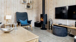A cozy living room with a wooden interior, a black wood-burning stove, a comfortable armchair, a flat-screen TV, and a dining area with a table and chairs. - 5 Star Holiday Home in Storvorde (Storvorde)