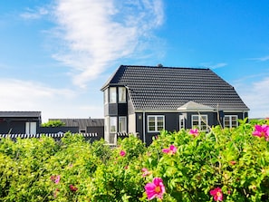 A house with a dark roof and white windows, surrounded by lush green foliage and vibrant pink flowers.