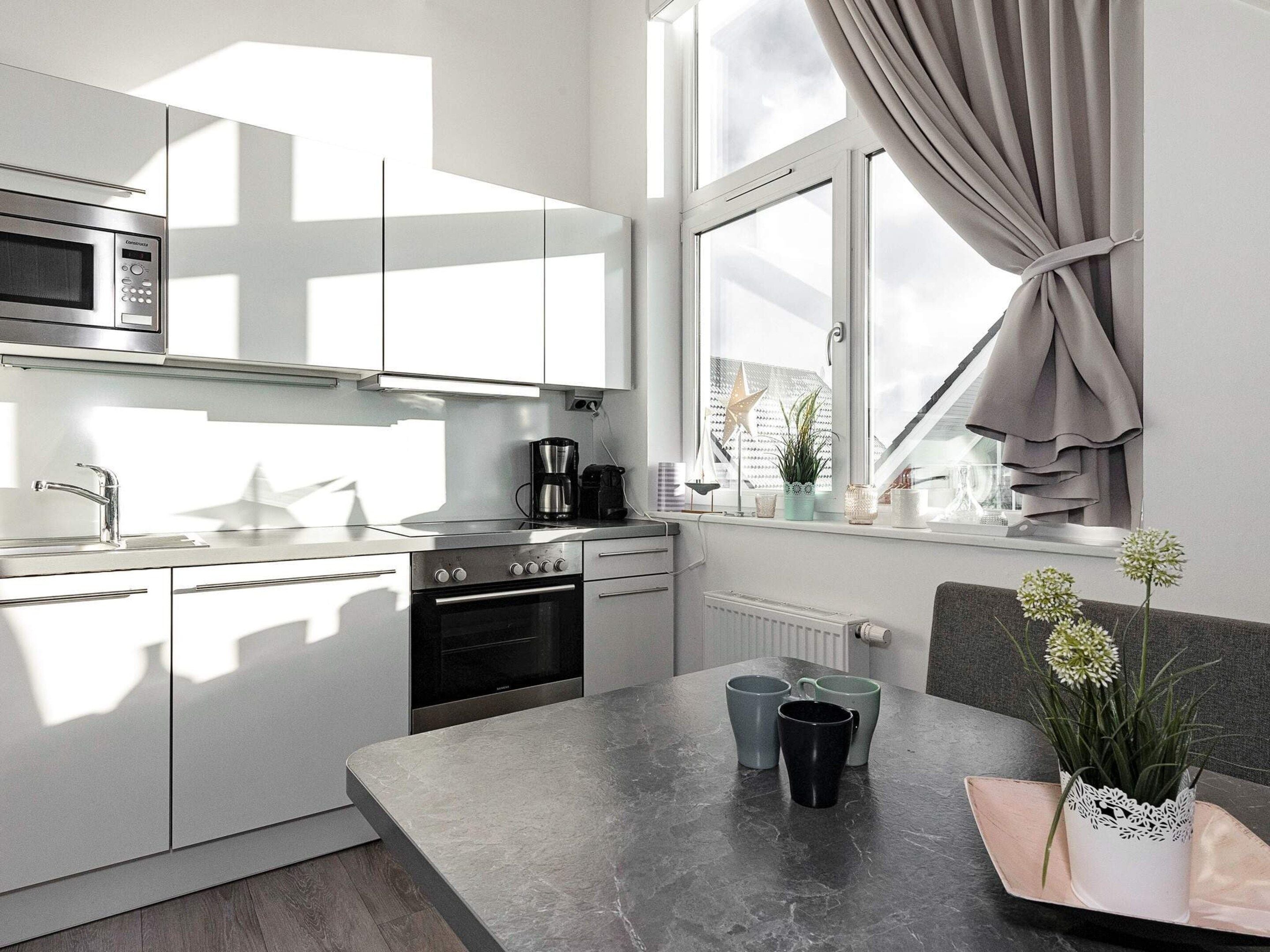 A modern kitchen with a built-in microwave, stove, and sink. There is a window with curtains, a plant on the windowsill, and a table with cups and a vase.