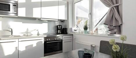 A modern kitchen with a built-in microwave, stove, and sink. There is a window with curtains, a plant on the windowsill, and a table with cups and a vase.