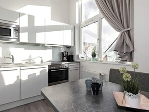 Une cuisine moderne équipée d’un four à micro-ondes, d’une cuisinière et d’un évier encastrés. On y trouve une fenêtre avec des rideaux, une plante sur le rebord de la fenêtre, ainsi qu’une table avec des tasses et un vase.