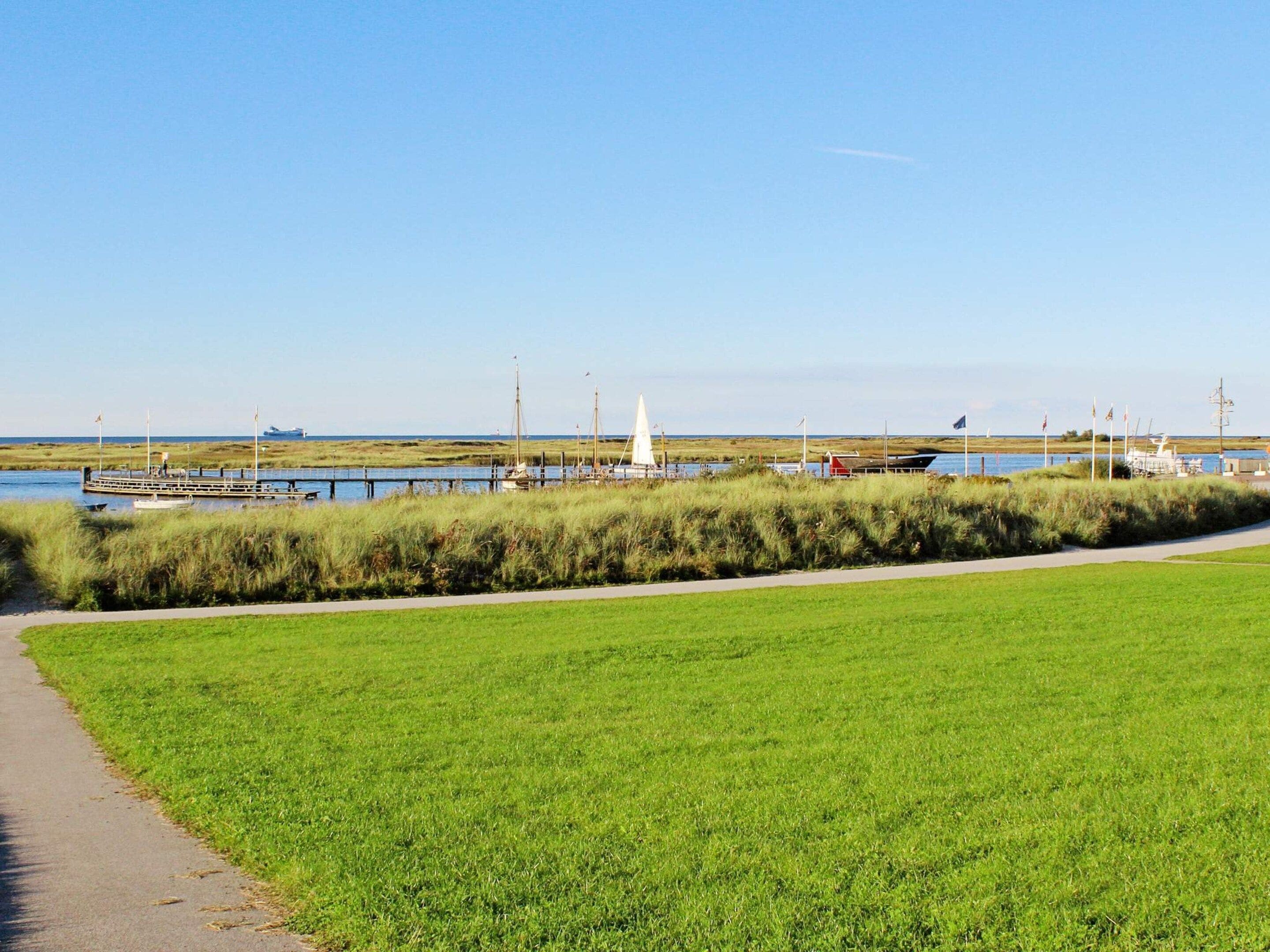 Een grasveld met een pad dat leidt naar een waterpartij, boten en een zeilboot die aan de oever liggen.