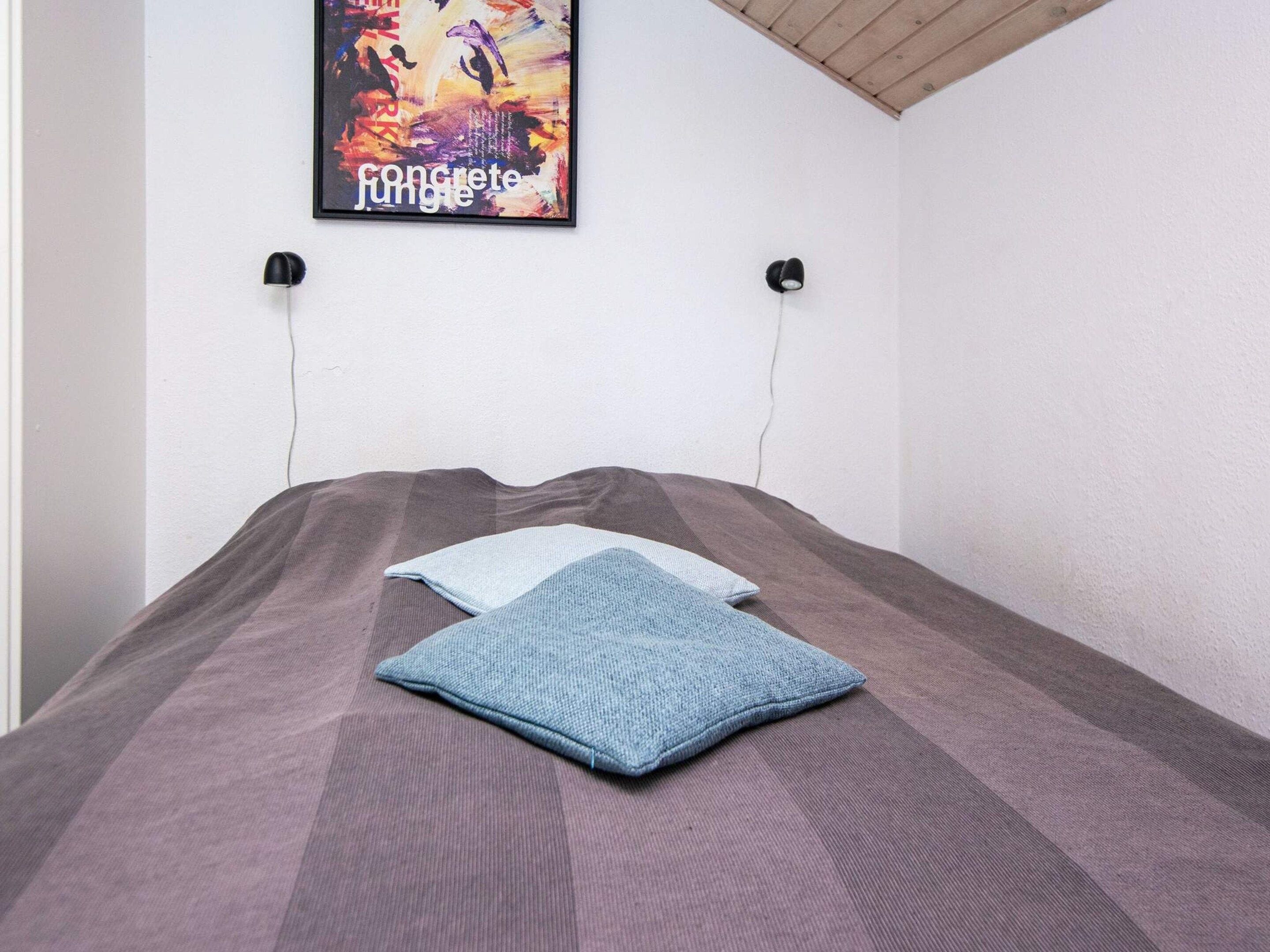 A bedroom with a bed, two pillows, a wall-mounted light, and a poster.