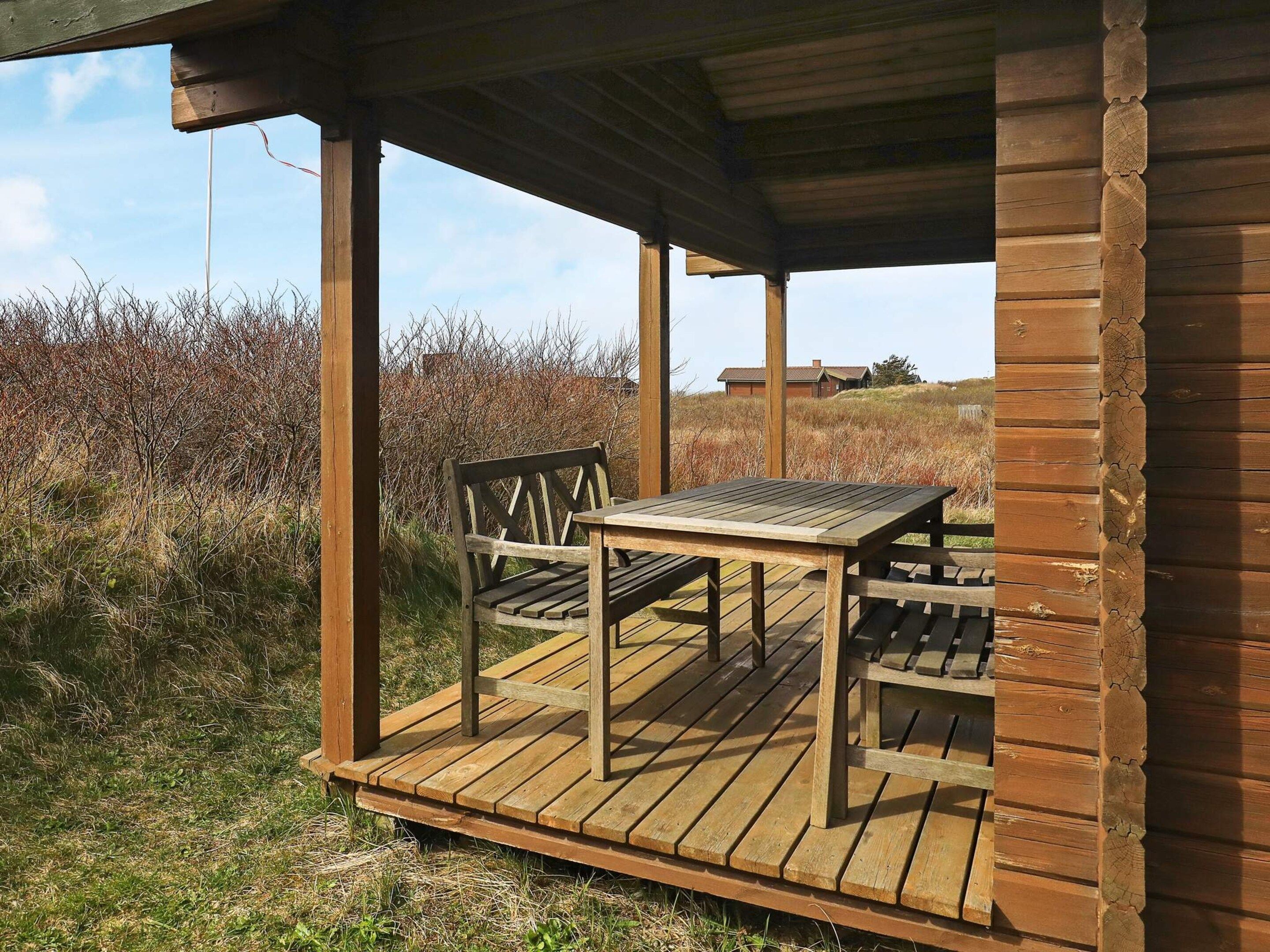 A wooden deck with a table and chairs, surrounded by natural vegetation and a distant building.