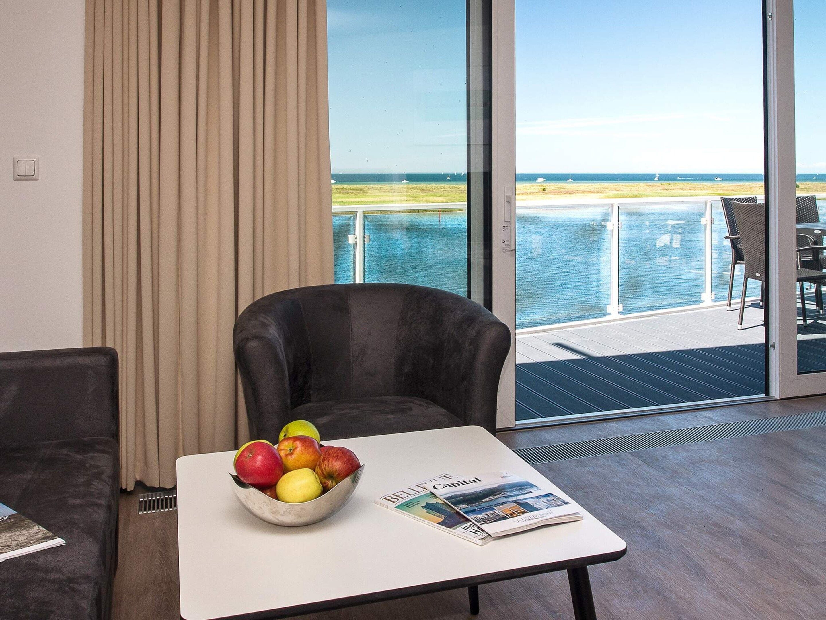 A room with a view of the sea, a table with a bowl of fruit, and a magazine.