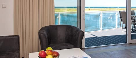 A room with a view of the sea, a table with a bowl of fruit, and a magazine.