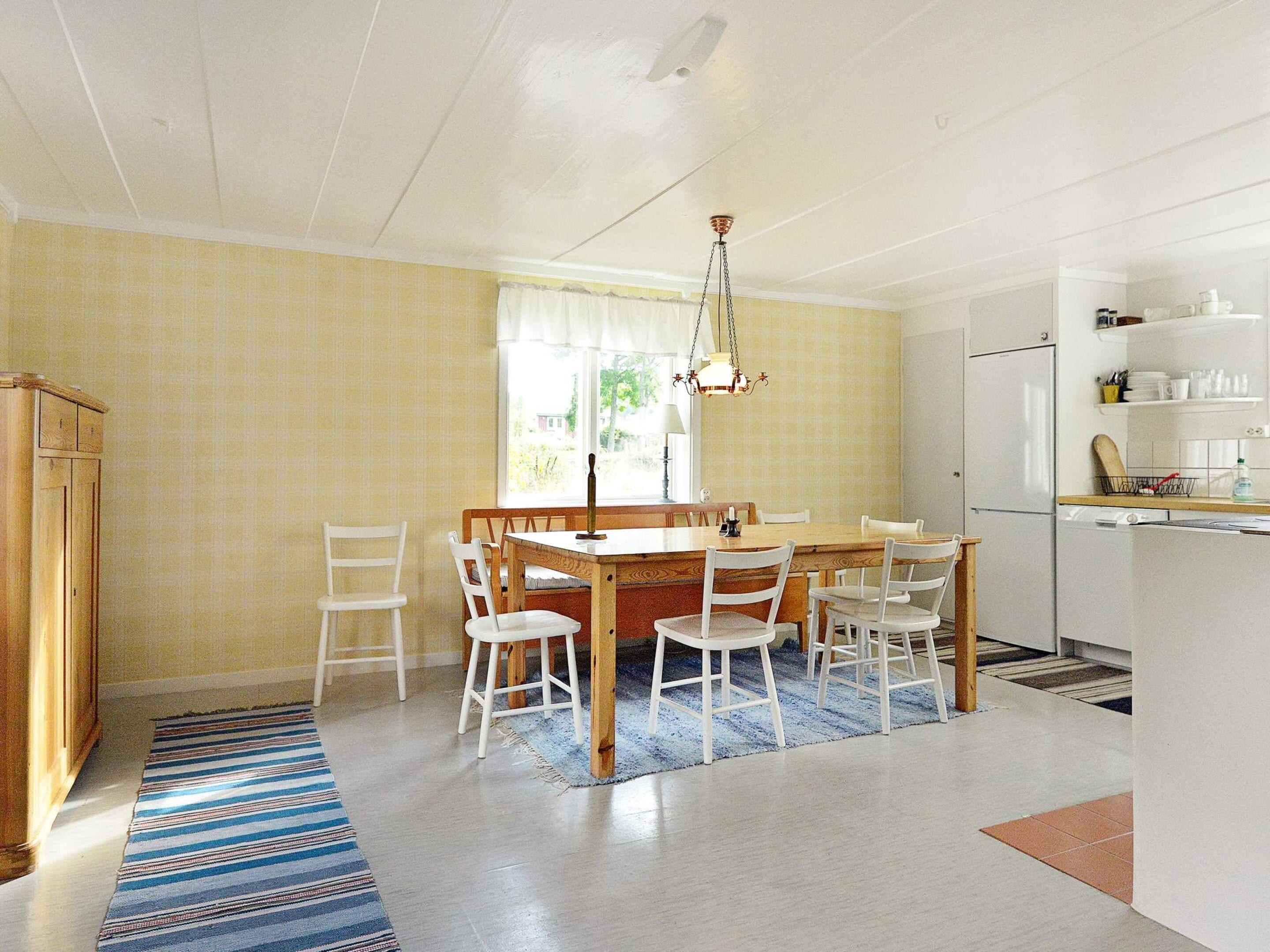 A dining area with a wooden table and white chairs, a hanging light fixture, and a kitchen area with white cabinets and a countertop.