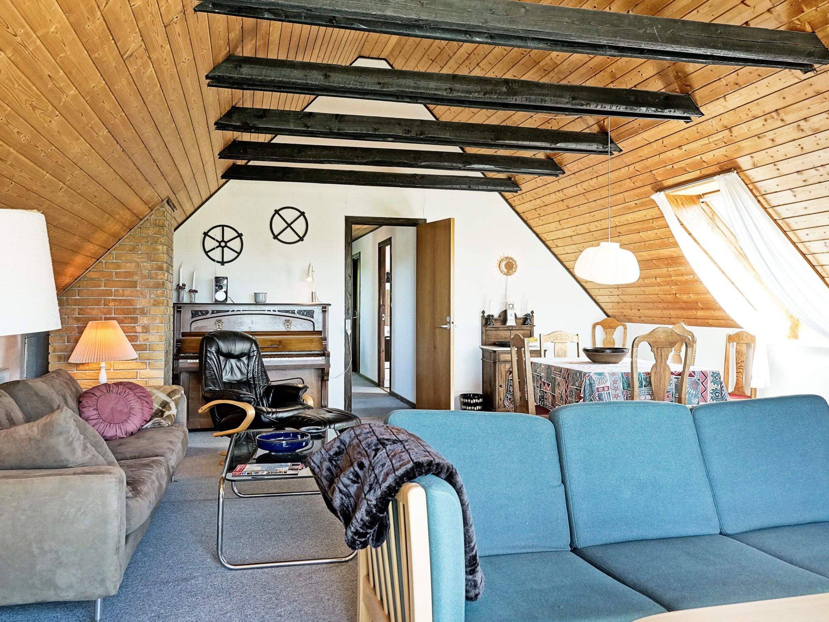 An attic room with a sloped ceiling, a piano, a dining table with chairs, a sofa, and a radiator.