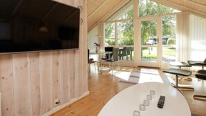 Ett modernt rum med ett träpanelat tak, stora fönster, en platt-TV, ett runt bord med fjärrkontroller och en matplats med stolar och bord.