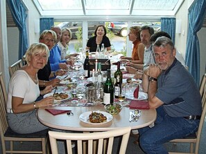 En gruppe mennesker sidder omkring et rundt bord med mad og drikke i et rum med et glasloft og gardiner.