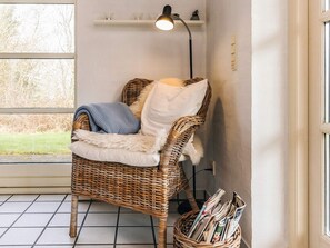 A wicker chair with a fur throw and pillows, a floor lamp, and a basket of newspapers on a tiled floor near a window. - 4 Star Holiday Home in Fur (Fur)