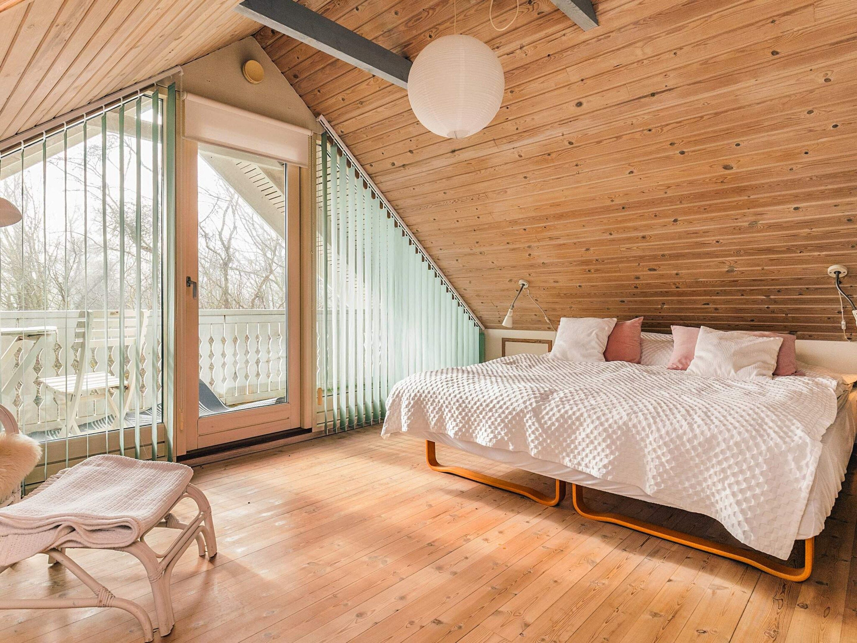 Un dormitorio con techo inclinado, una cama con colcha blanca, un banco y vistas a través de un ventanal.
