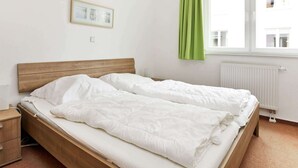 A single bed with a wooden headboard, white bedding, a small bedside table, and a window with green curtains. - Holiday Home in Grossenbrode (Grossenbrode)