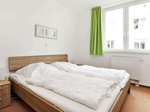 Ein Einzelbett mit Holz Kopfteil, weißer Bettwäsche, einem kleinen Nachttisch und einem Fenster mit grünen Vorhängen.