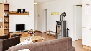 A modern living room with a sofa, a TV, a wooden bookshelf, and a small table with a vase of flowers. - Holiday Home in Grossenbrode (Grossenbrode)
