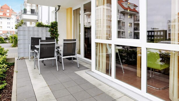 A modern apartment with a patio area featuring a table and chairs, large windows, and a view of neighboring buildings. - Holiday Home in Grossenbrode (Grossenbrode)