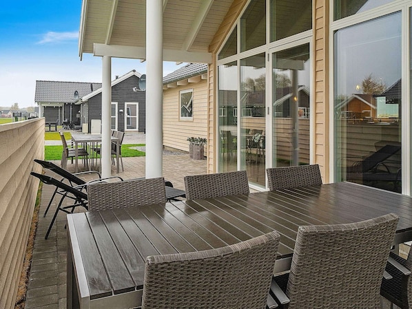 A covered patio with a wooden table and wicker chairs, large glass sliding doors, and a view of neighboring houses.