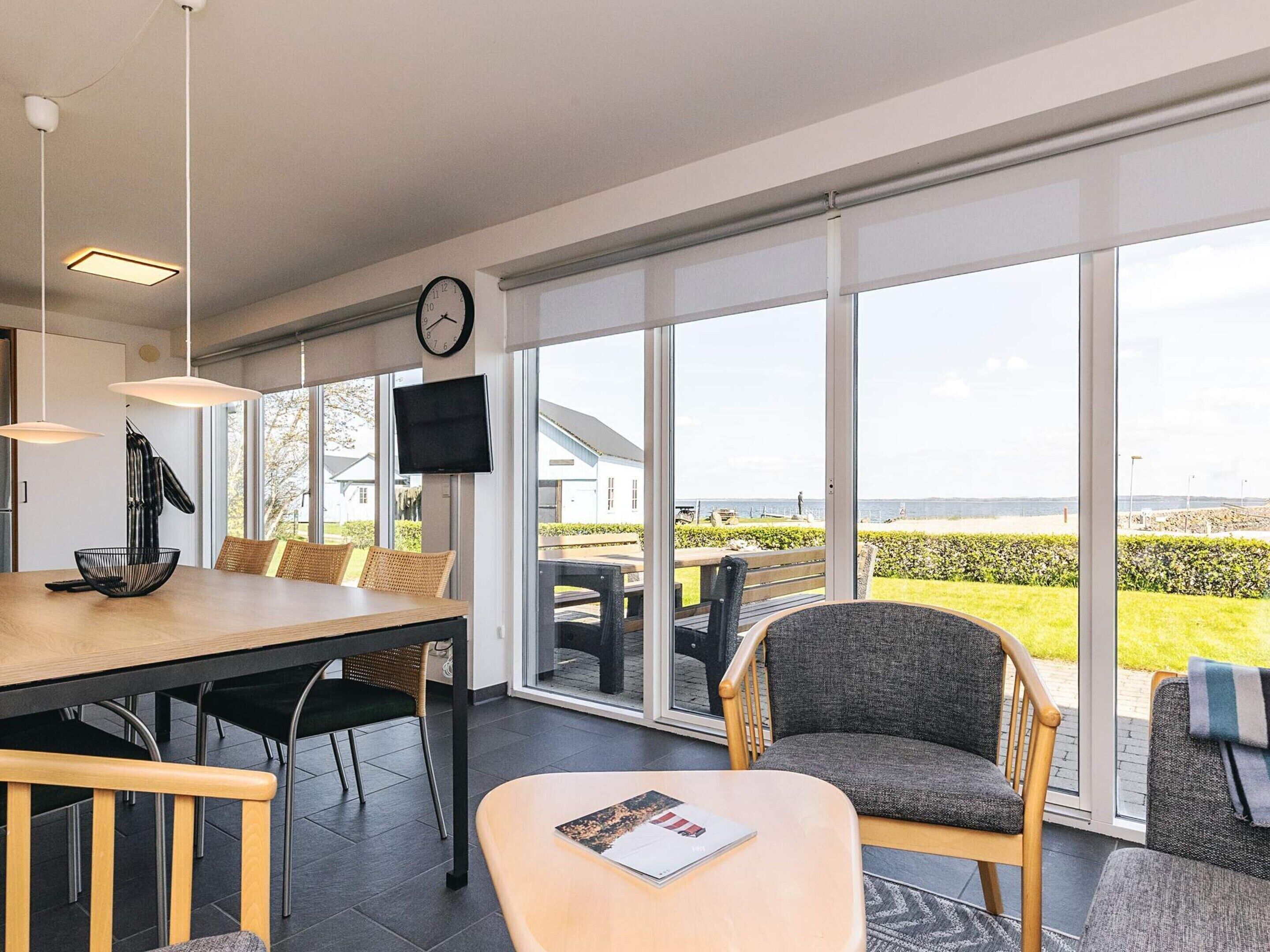 Un espace repas moderne avec une table en bois, des chaises, un téléviseur et une grande fenêtre donnant sur la mer.