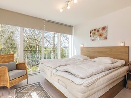 Un dormitorio con una cama grande, una silla de mimbre, vistas a árboles por la ventana y un cuadro en la pared.