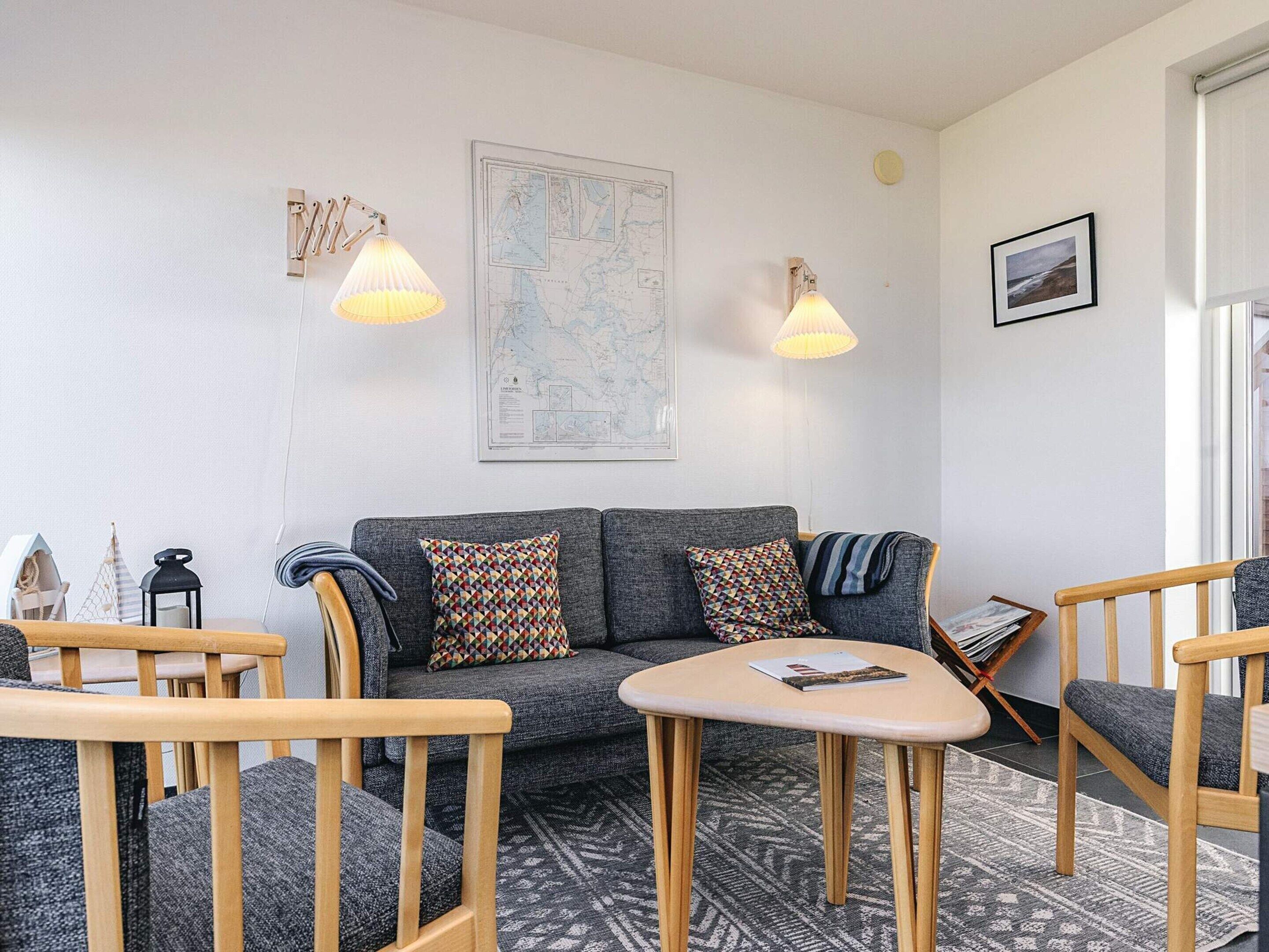 A cozy living room with a grey sofa, wooden chairs, a small table, and two wall-mounted lamps.