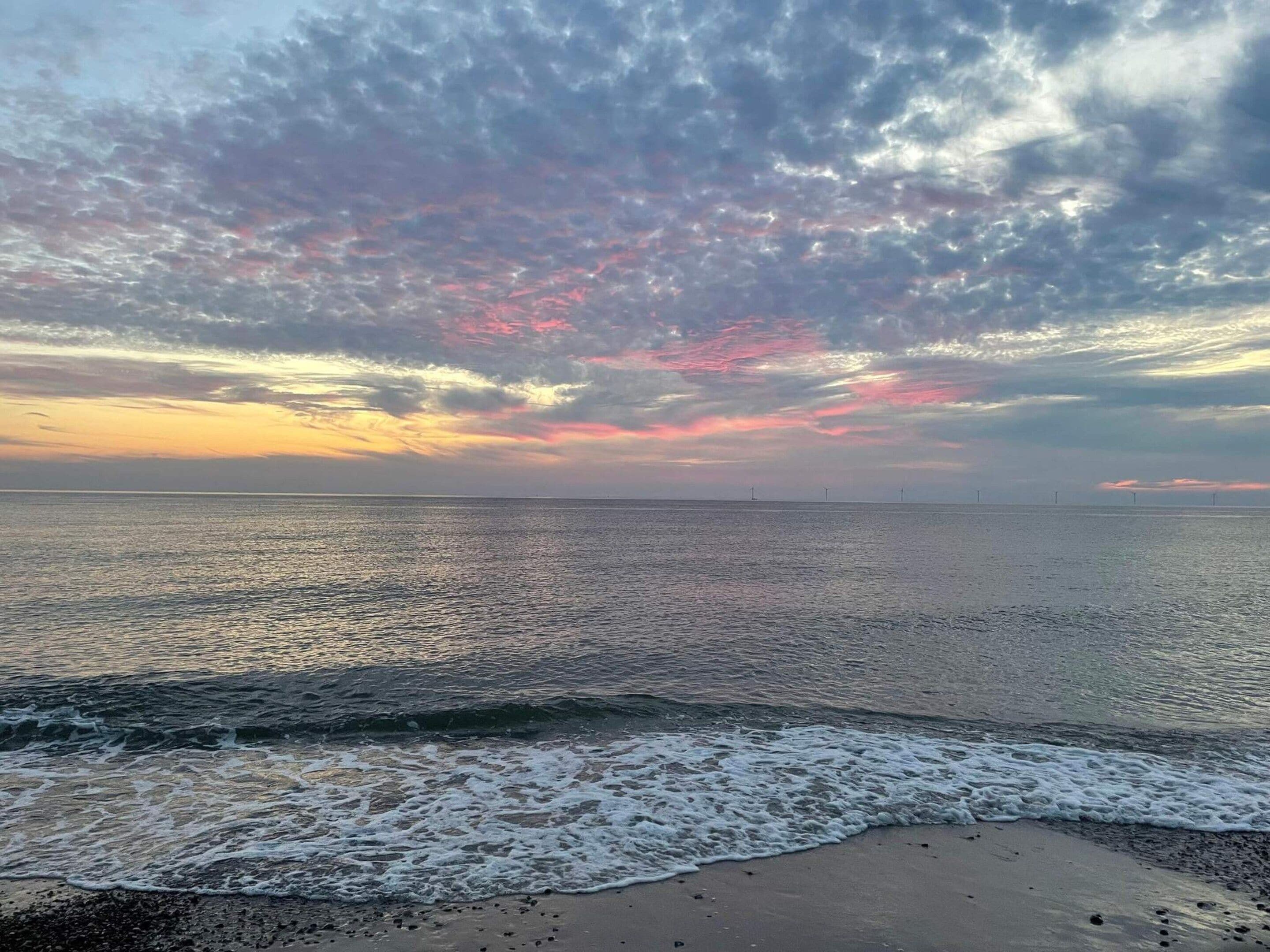 Una playa con olas, cielo despejado y aerogeneradores a lo lejos.