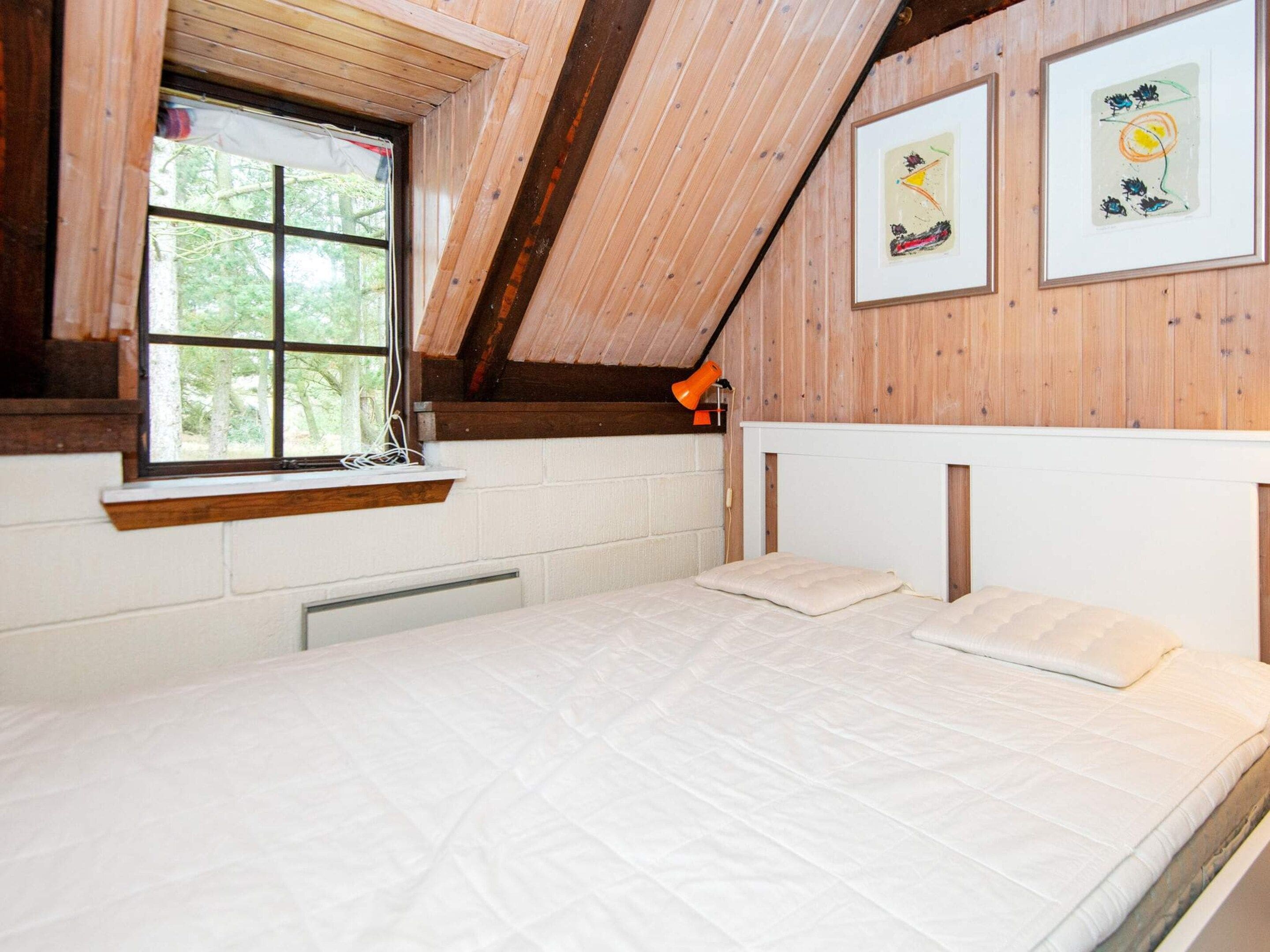 Una habitación con techo inclinado, paredes de madera, un ventanal, una cama y dos obras de arte enmarcadas.