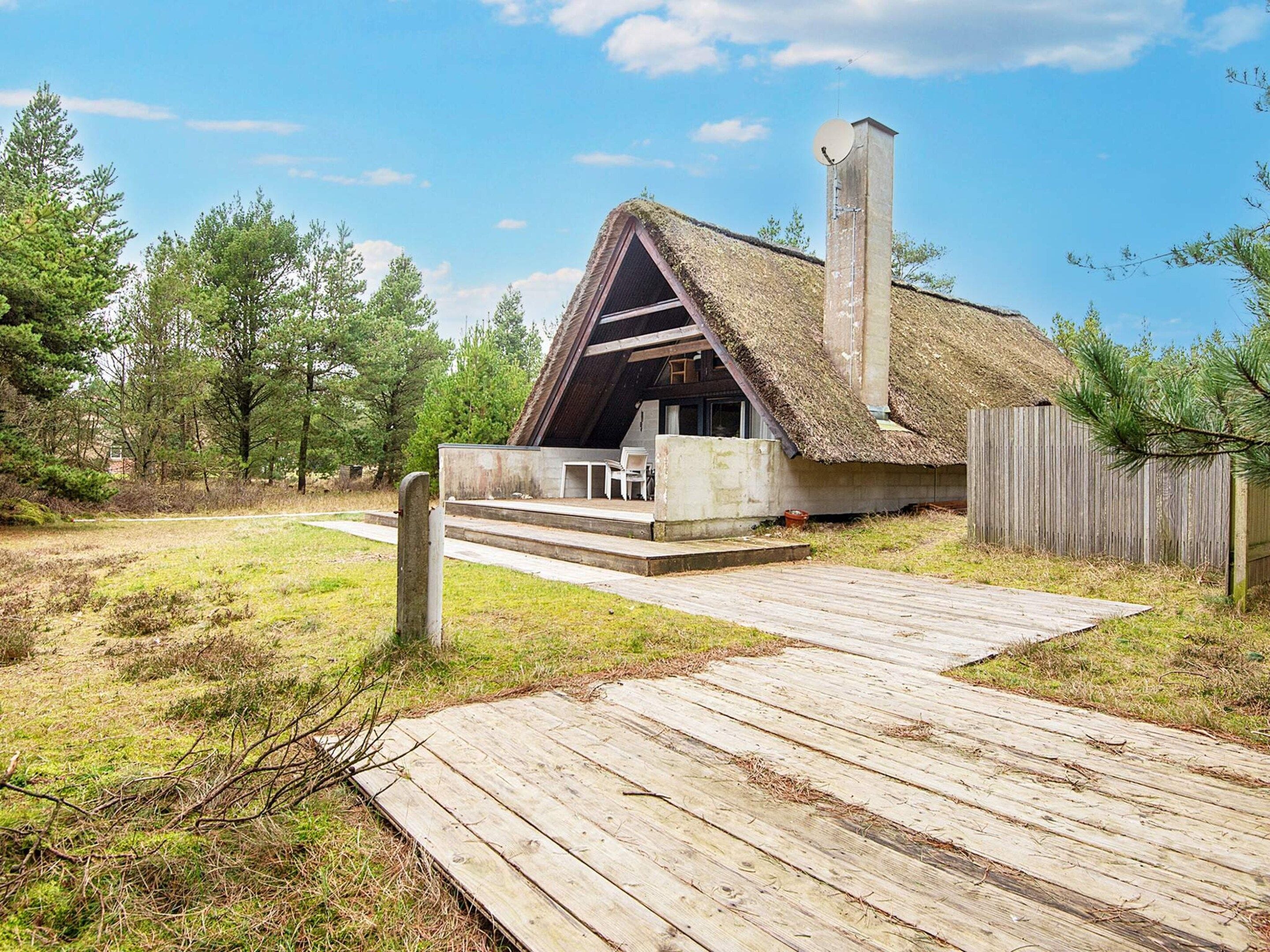 Ein rustikales Blockhaus mit Strohdach, Schornstein und einer Holzterrasse, die zum Eingang führt.