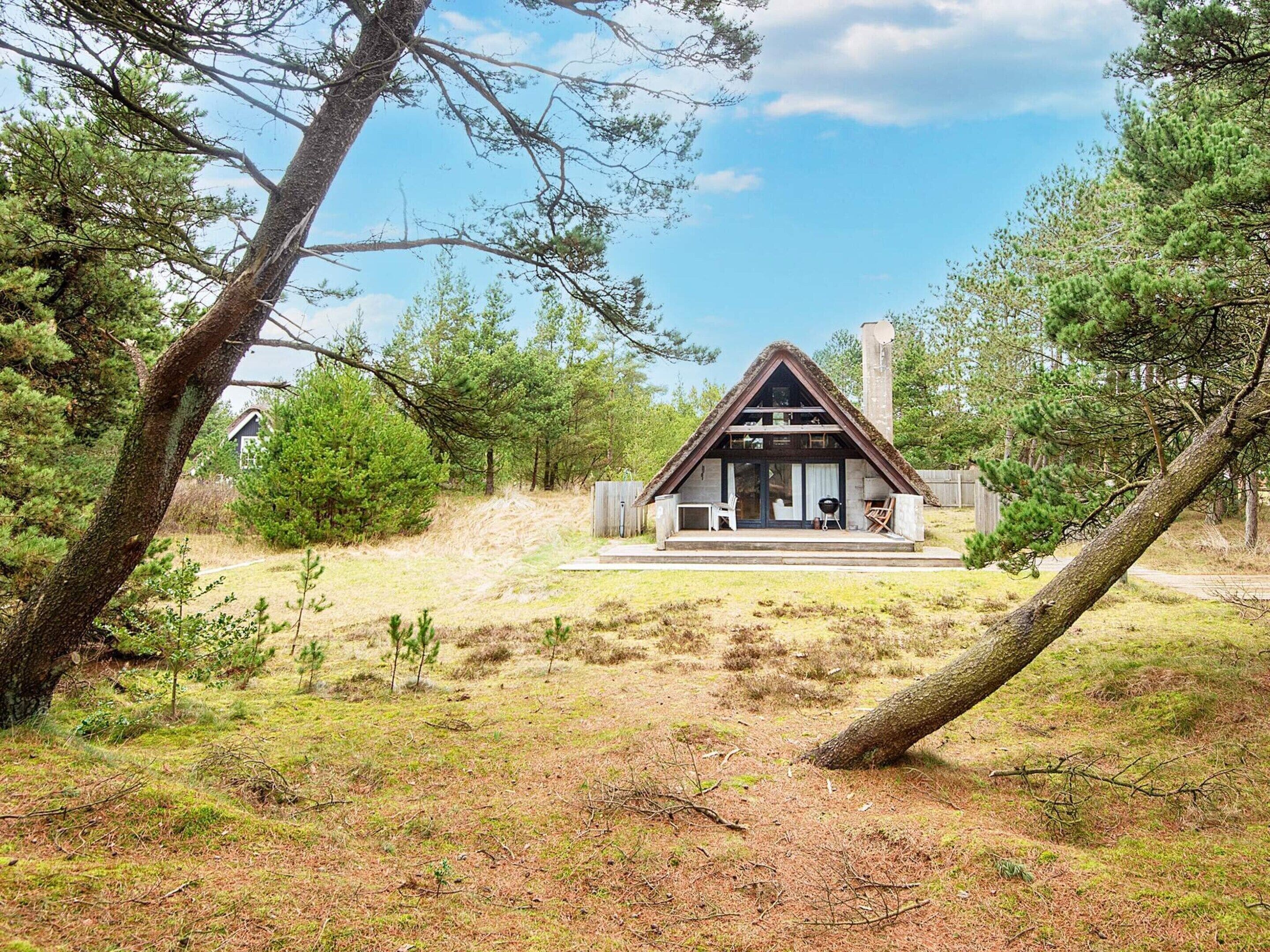 A cabin with a thatched roof, a porch, and a chimney, surrounded by trees and a grassy area.