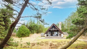 A cabin with a thatched roof, a porch, and a chimney, surrounded by trees and a grassy area.