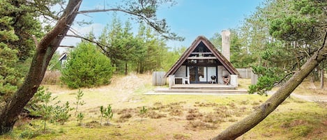 A cabin with a thatched roof, a porch, and a chimney, surrounded by trees and a grassy area.
