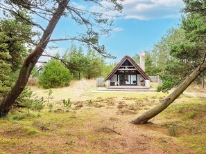 A cabin with a thatched roof, a porch, and a chimney, surrounded by trees and a grassy area.
