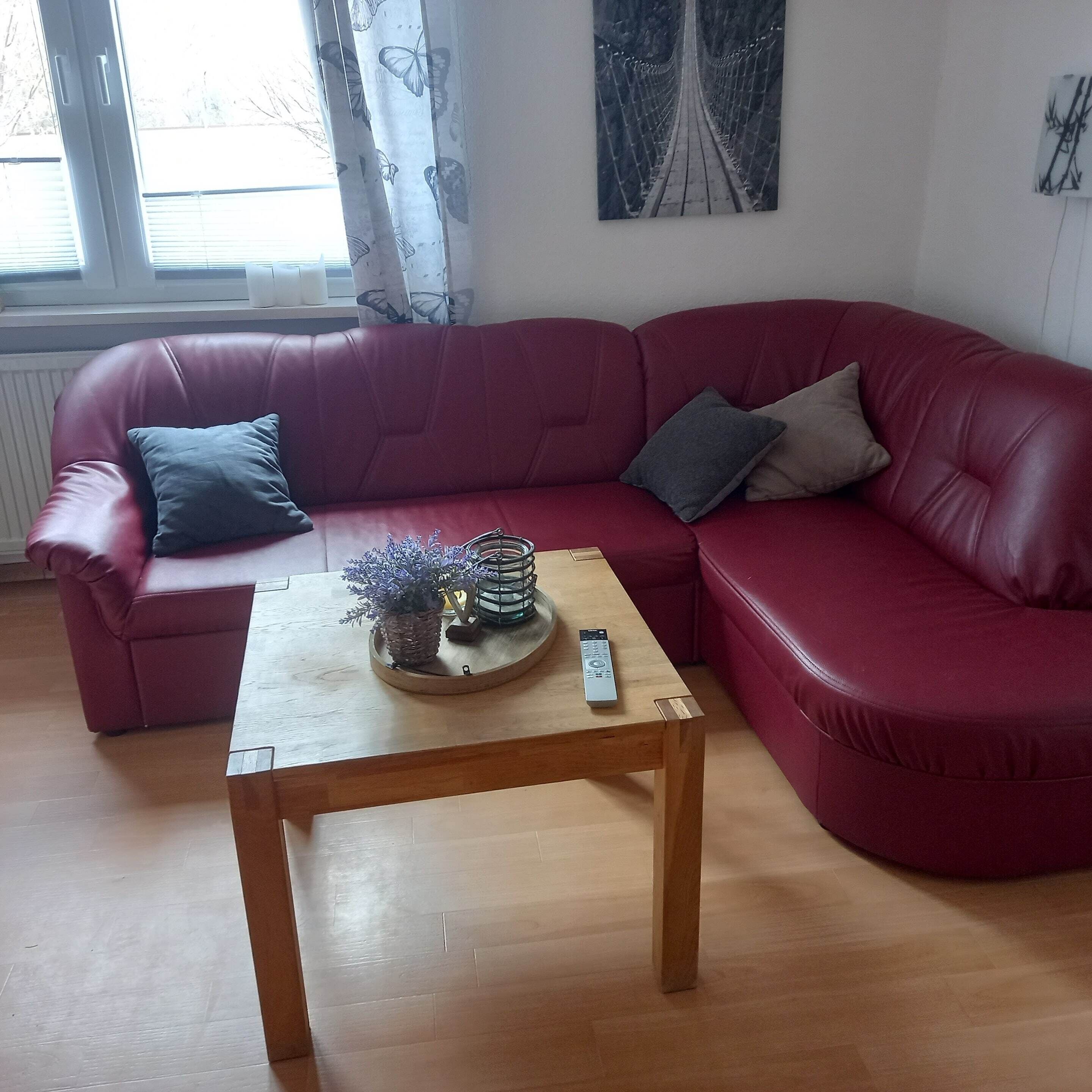 A red leather sofa with cushions, a wooden coffee table with decorative items, and a window with curtains.